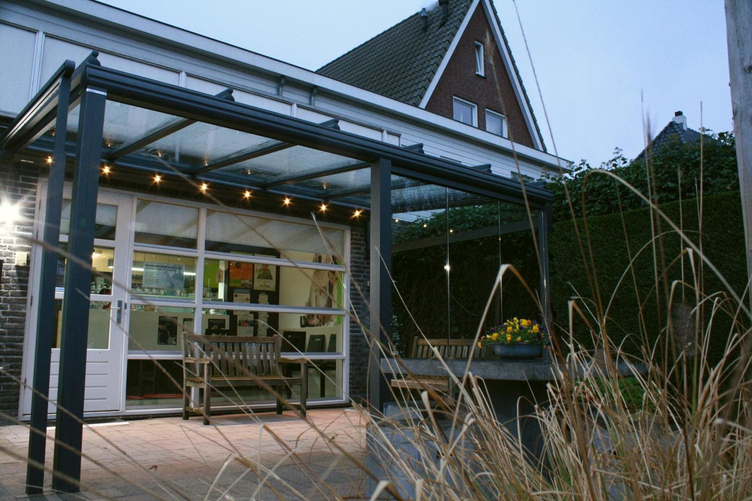 De terrasoverkapping aan de praktijk bij schemer, met verlichting en banken voor de ingang